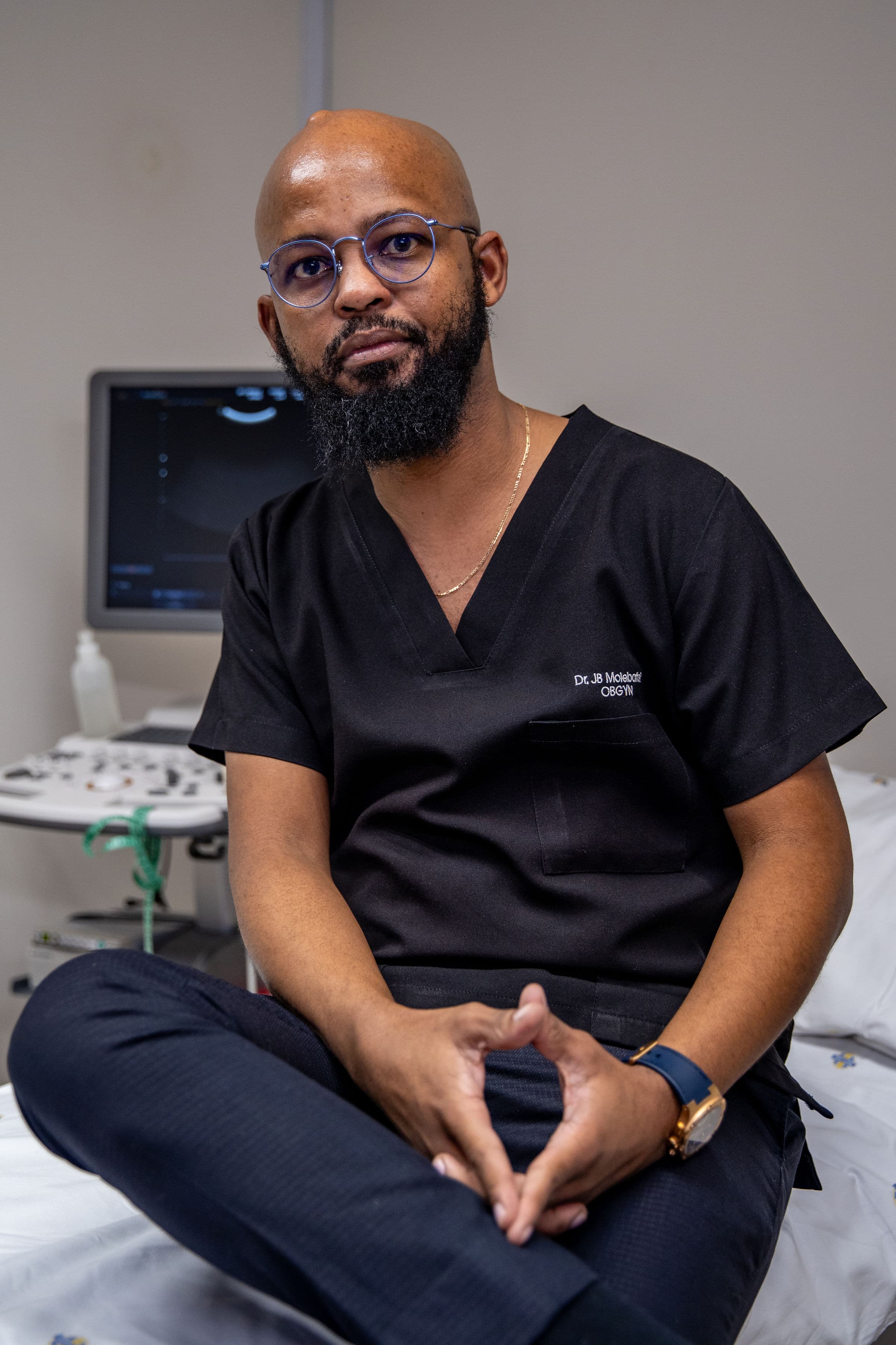 Bearded OBGYN in black scrubs sits on a medical bed near an ultrasound machine.