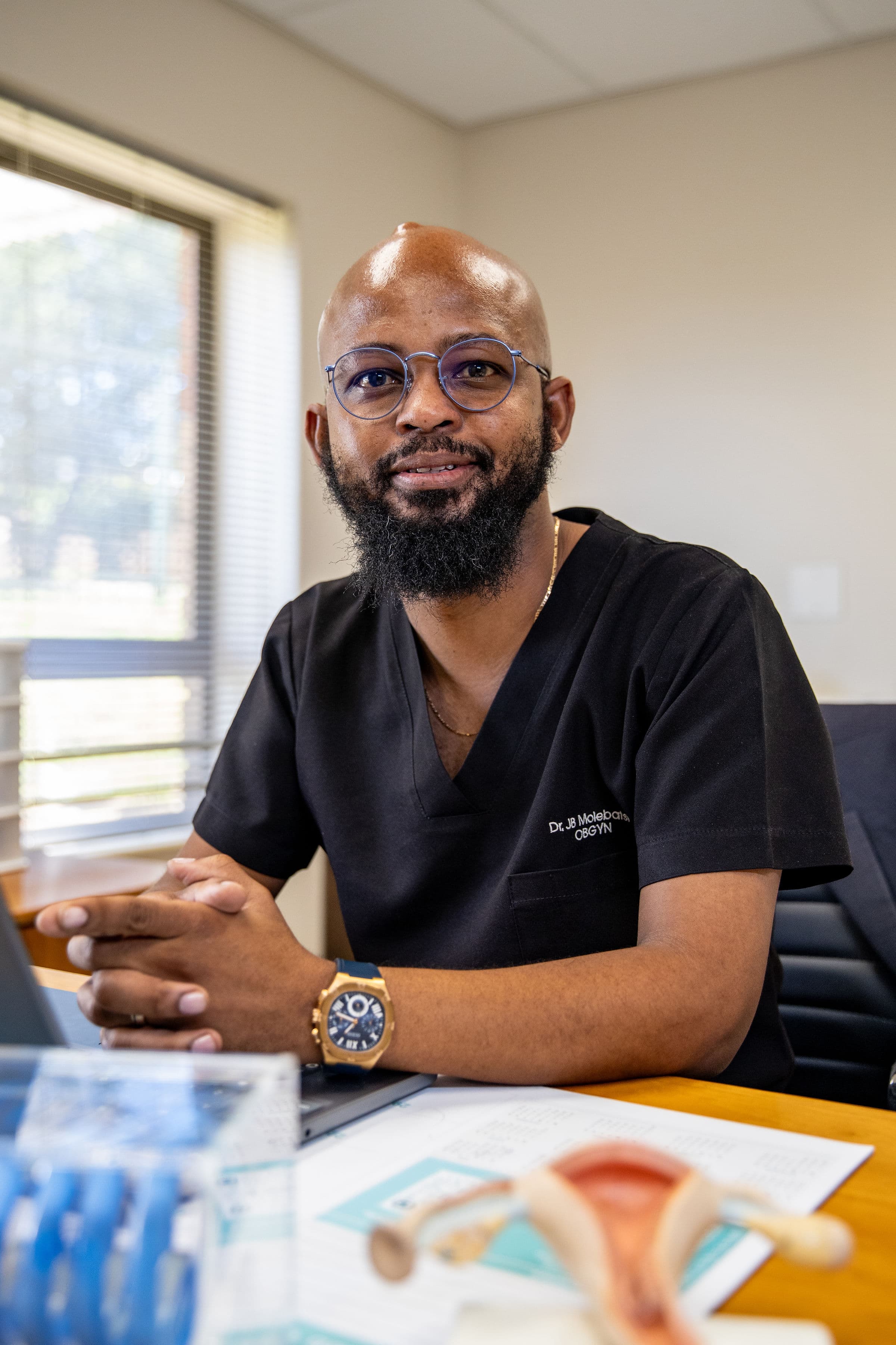 Dr. JB Molebatsi, an OBGYN, sits at his desk wearing black scrubs and glasses.