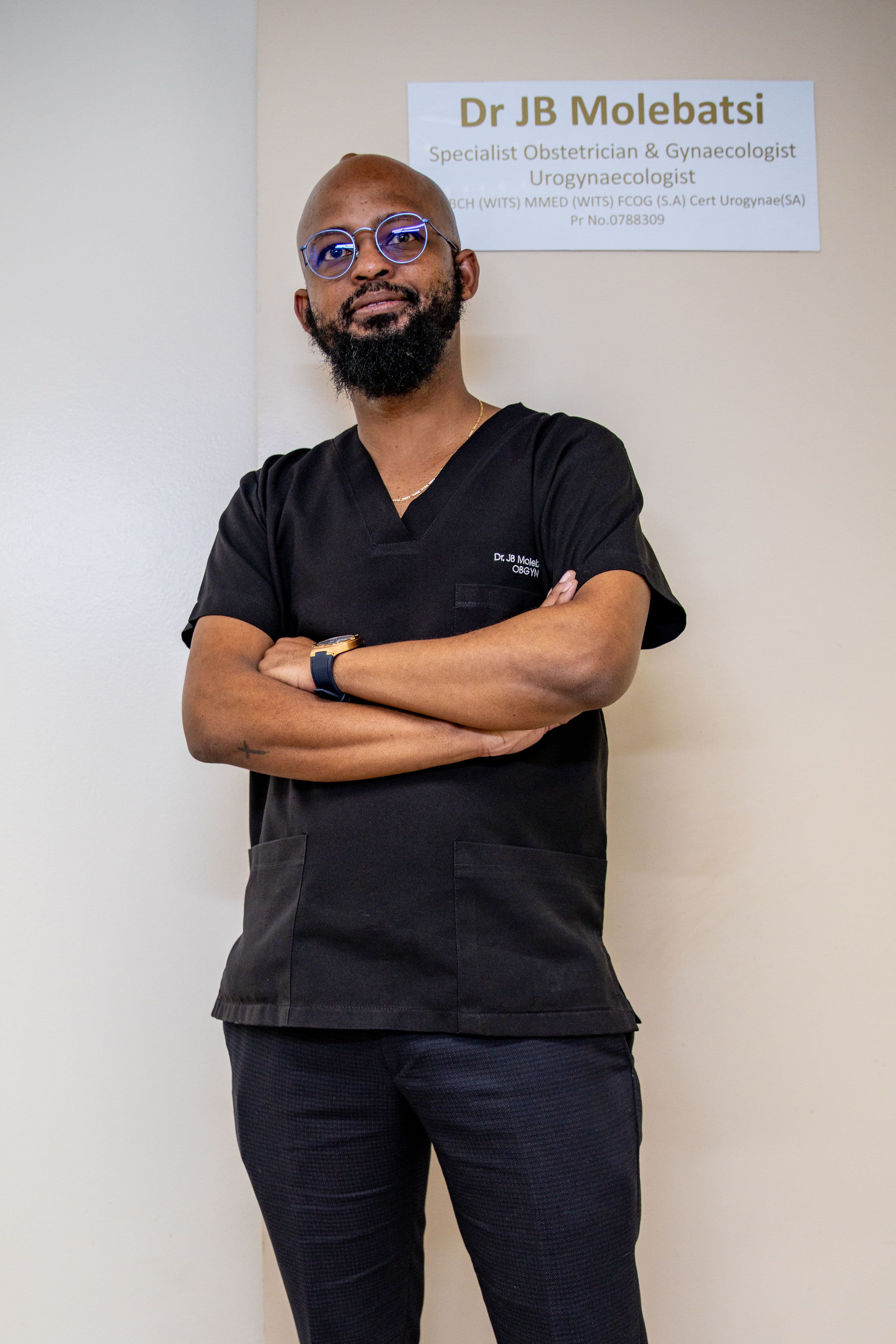 Bearded OBGYN Dr. JB Molebatsi posing in black scrubs before his office sign.